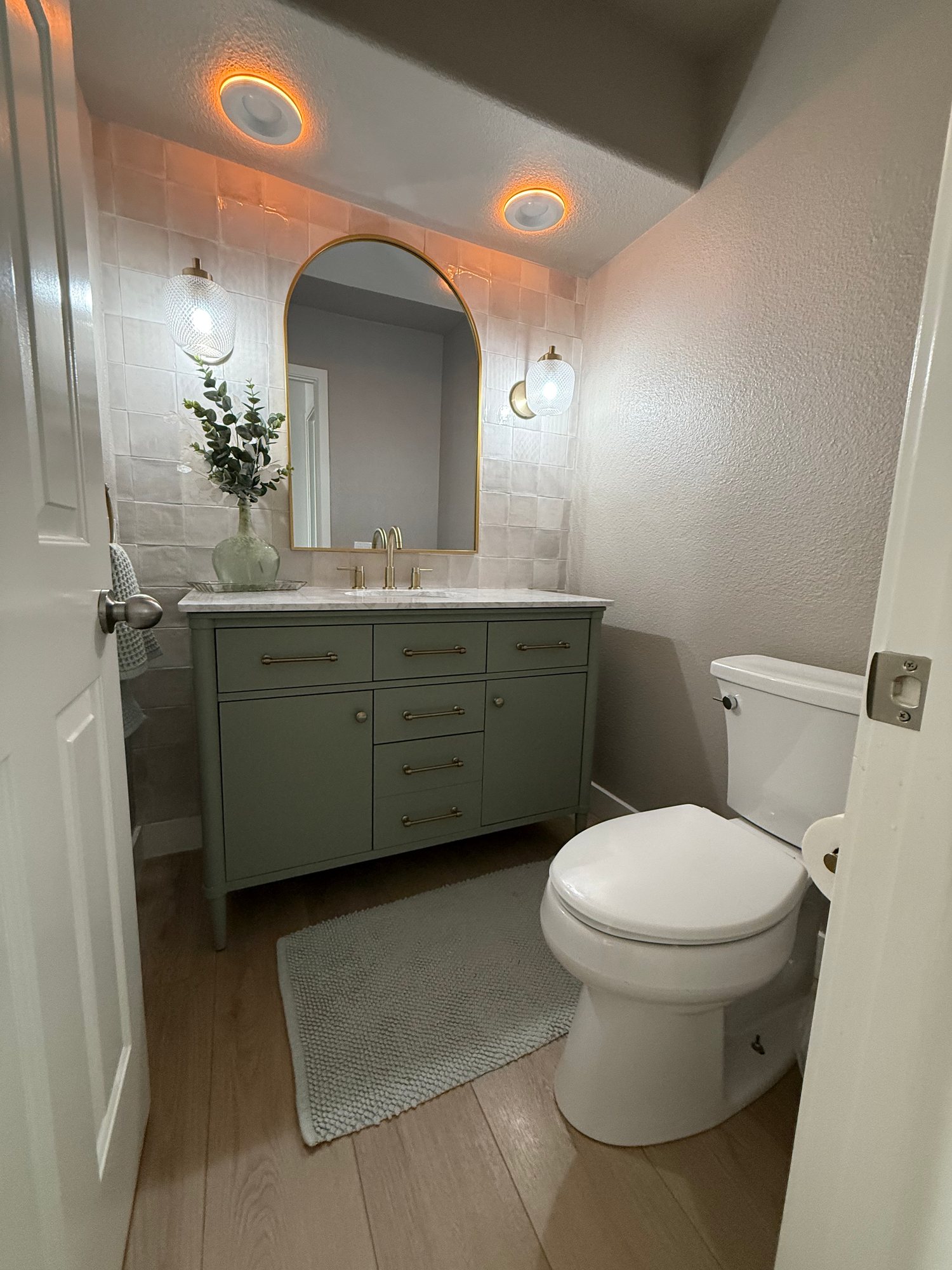 Powder room with a sage-green vanity, arched brass mirror, and warm accent lighting