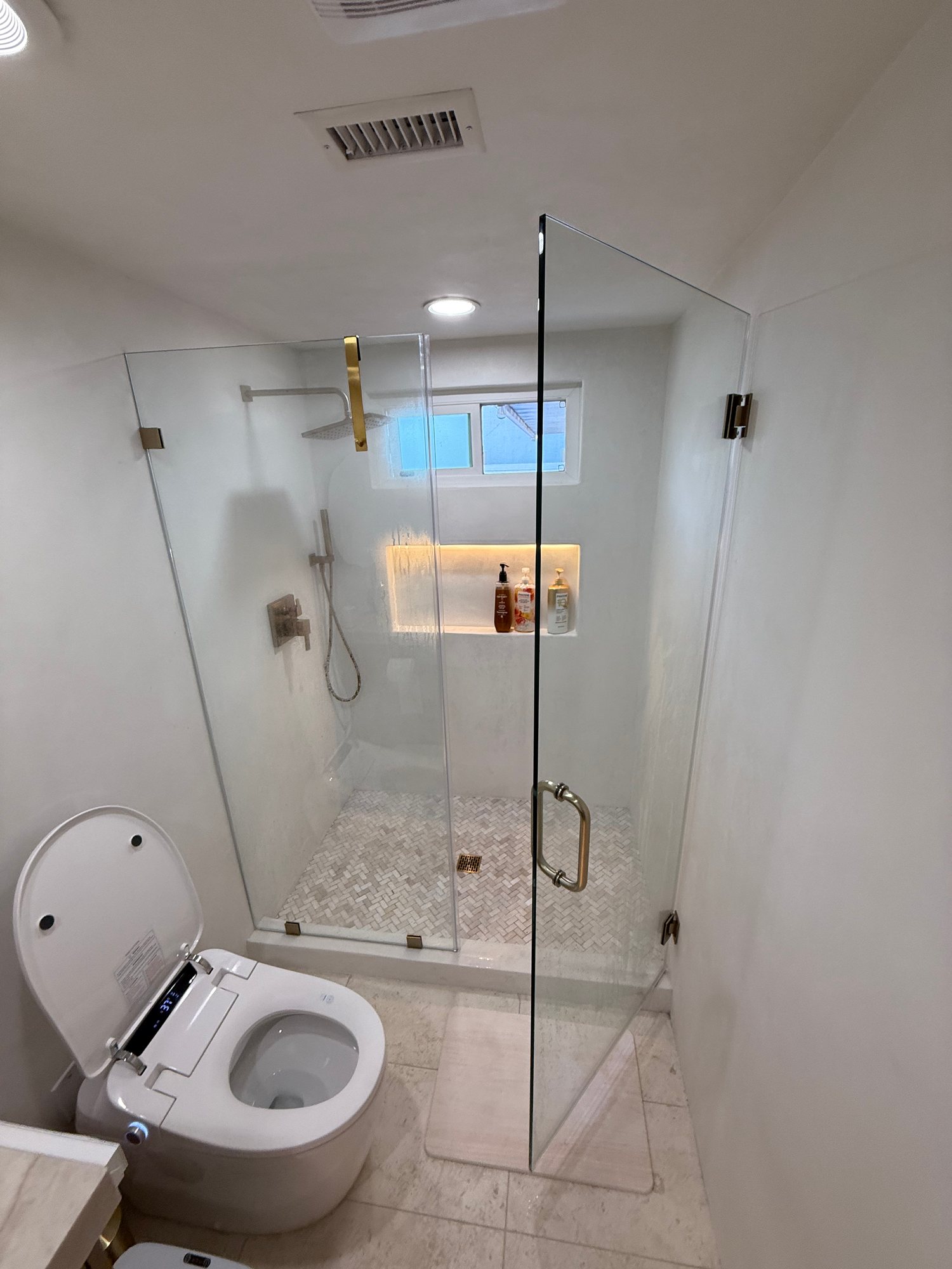 Glass walk-in shower with brass fixtures and a lit niche beside a smart toilet