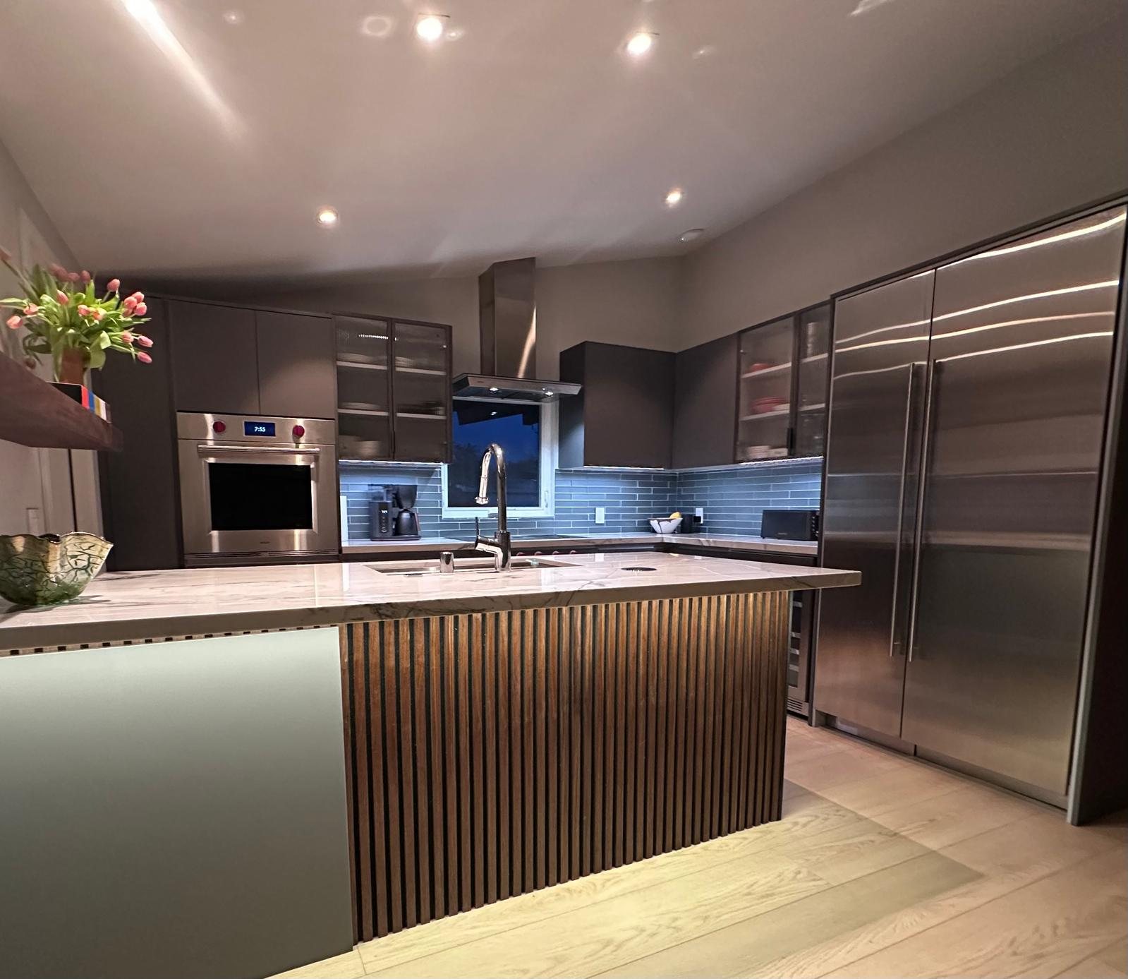 Kitchen with a wood-slat island, dark cabinetry, blue tile backsplash, and stainless built-in refrigerator